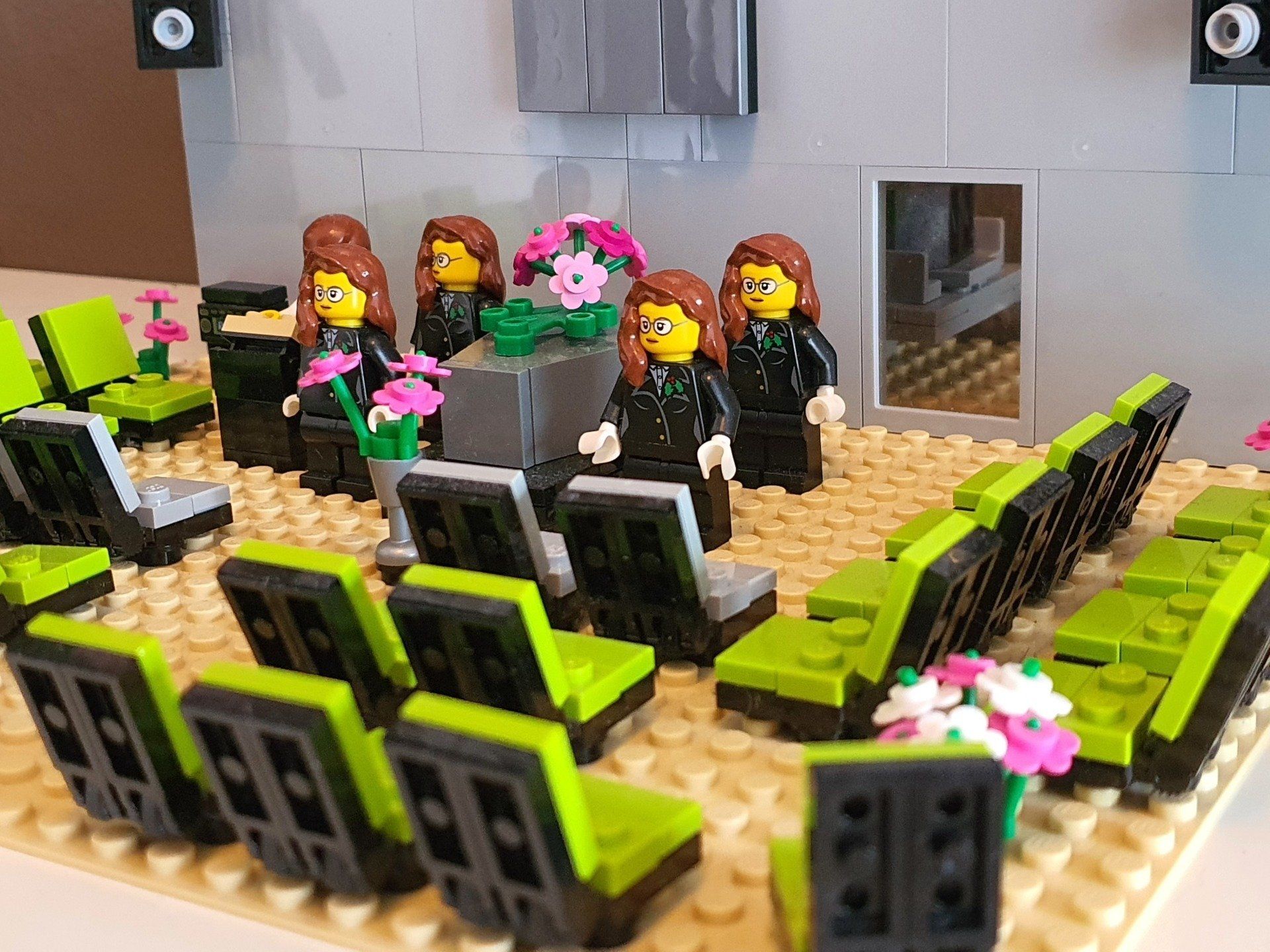 Een Lego-scène toont vier figuren die aan een tafel op een podium zitten, tegenover rijen groene en zwarte stoelen in een kamer.