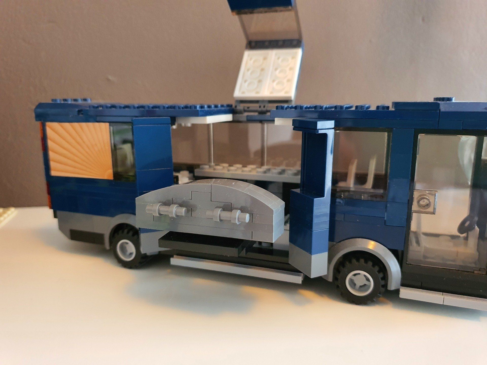 Een blauw-grijze LEGO-bus met een open zijpaneel en dak, die op een witte ondergrond tegen een effen muur staat.