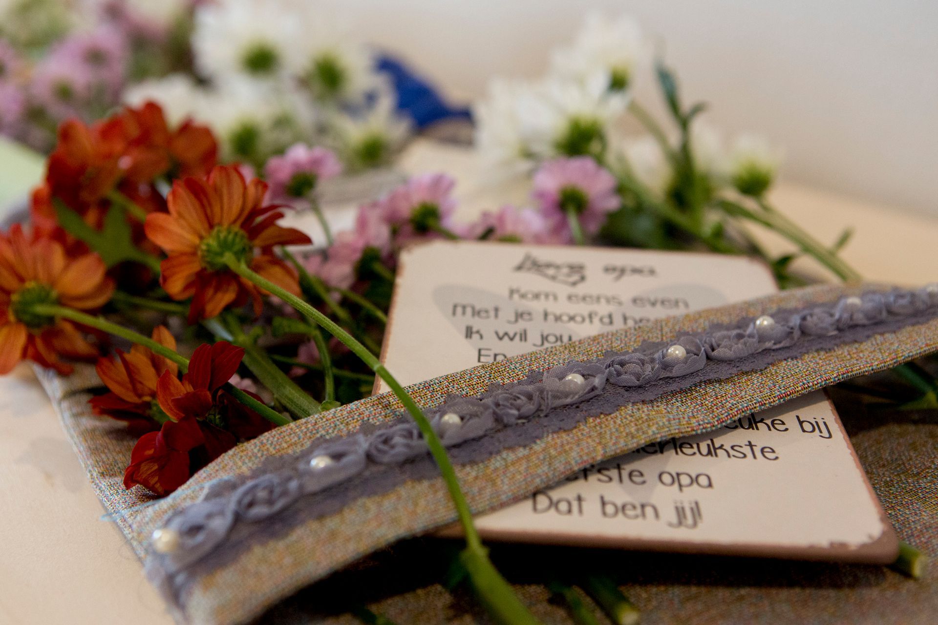 Een decoratieve kaart met Nederlandse tekst ligt tussen oranje en witte bloemen, omlijst door een grijs lint met kleine kraaltjes.