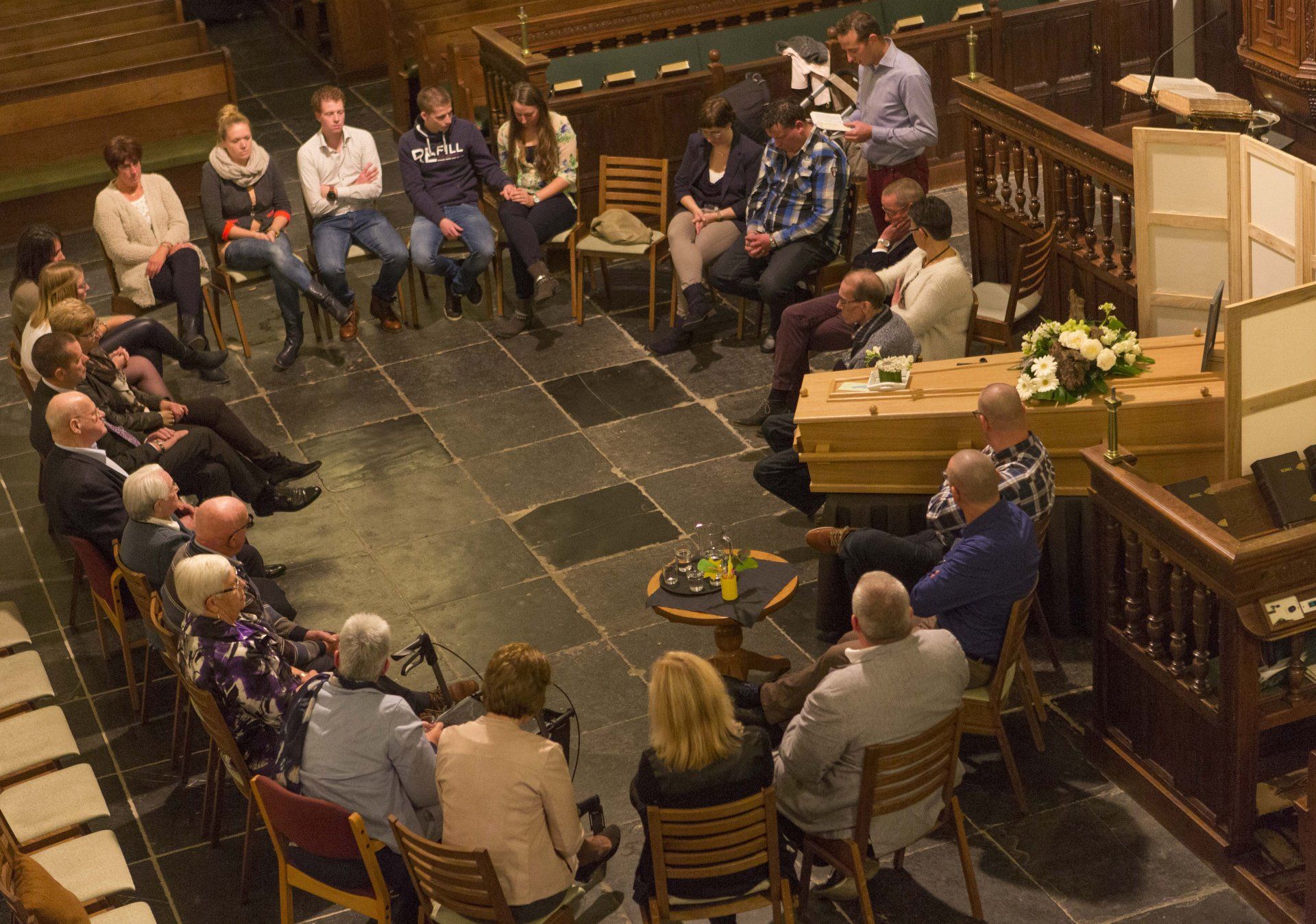 Een groep mensen zit in een grote cirkel op houten stoelen in een schemerig verlichte zaal, rondom een houten doodskist.