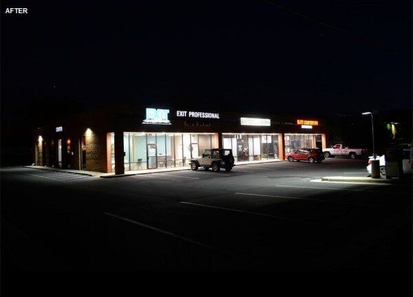 Night exterior of a strip mall lit up. Storefronts with signs and parked cars.
