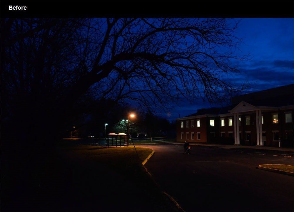 Dark night scene: a building with lit windows, a bare tree, and a person biking on a road.