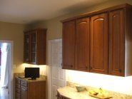 Kitchen cabinets: brown, with under-cabinet lighting.  Countertop with bowl and fruit, a TV on a lower cabinet.