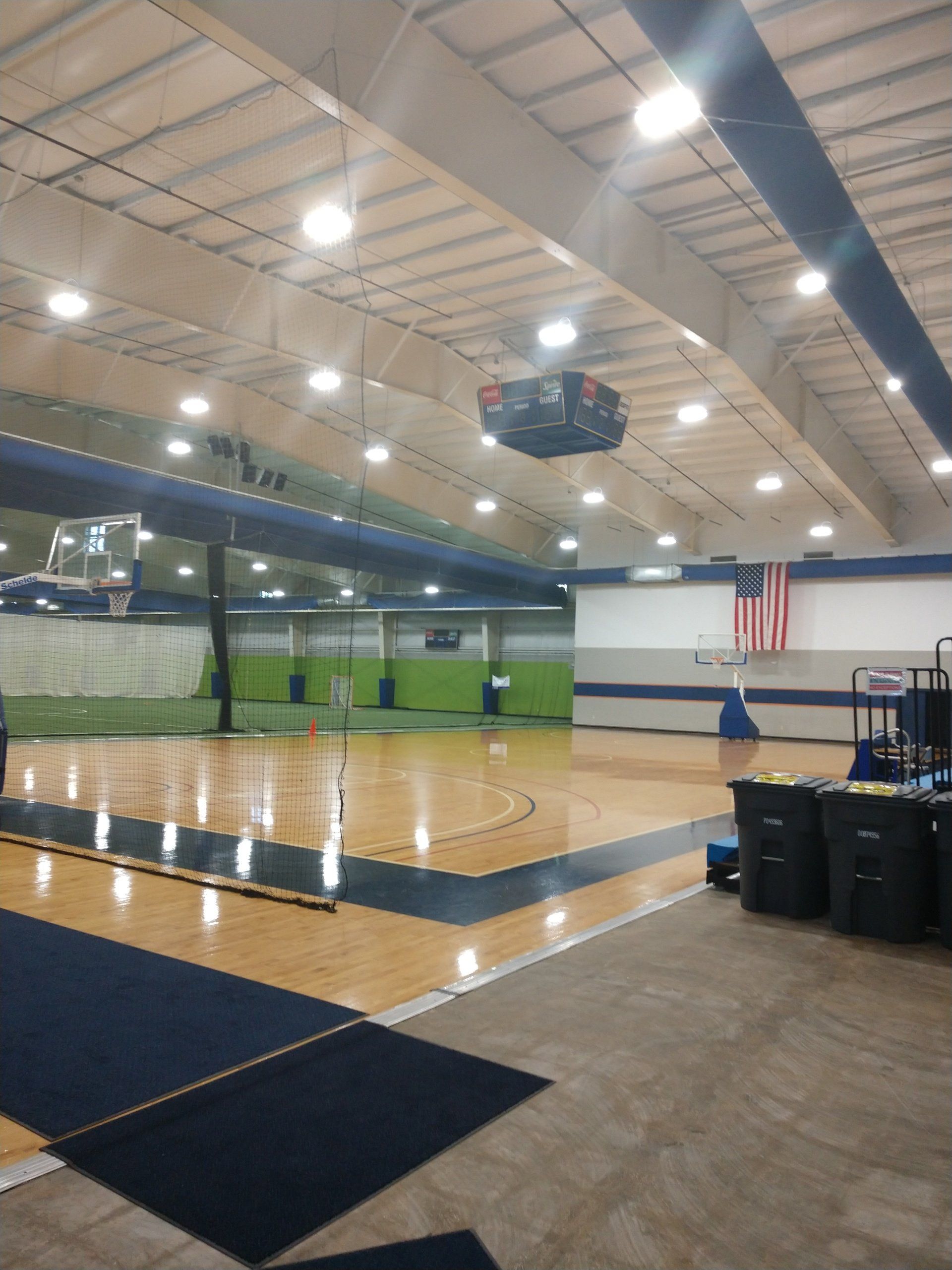 Basketball court in a gymnasium; wooden floor, blue accents, scoreboard, and American flag.