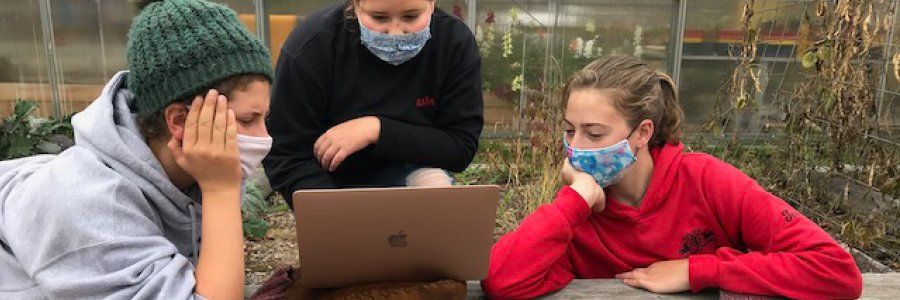 three adolescent students outside at a picnic table gathered around a laptop