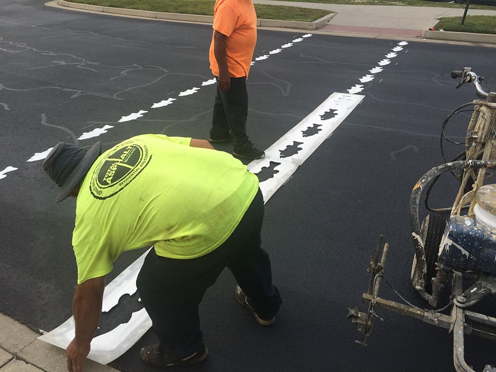 Two workers apply a stencil of small fish shapes to create a crosswalk on a freshly paved black asphalt road.