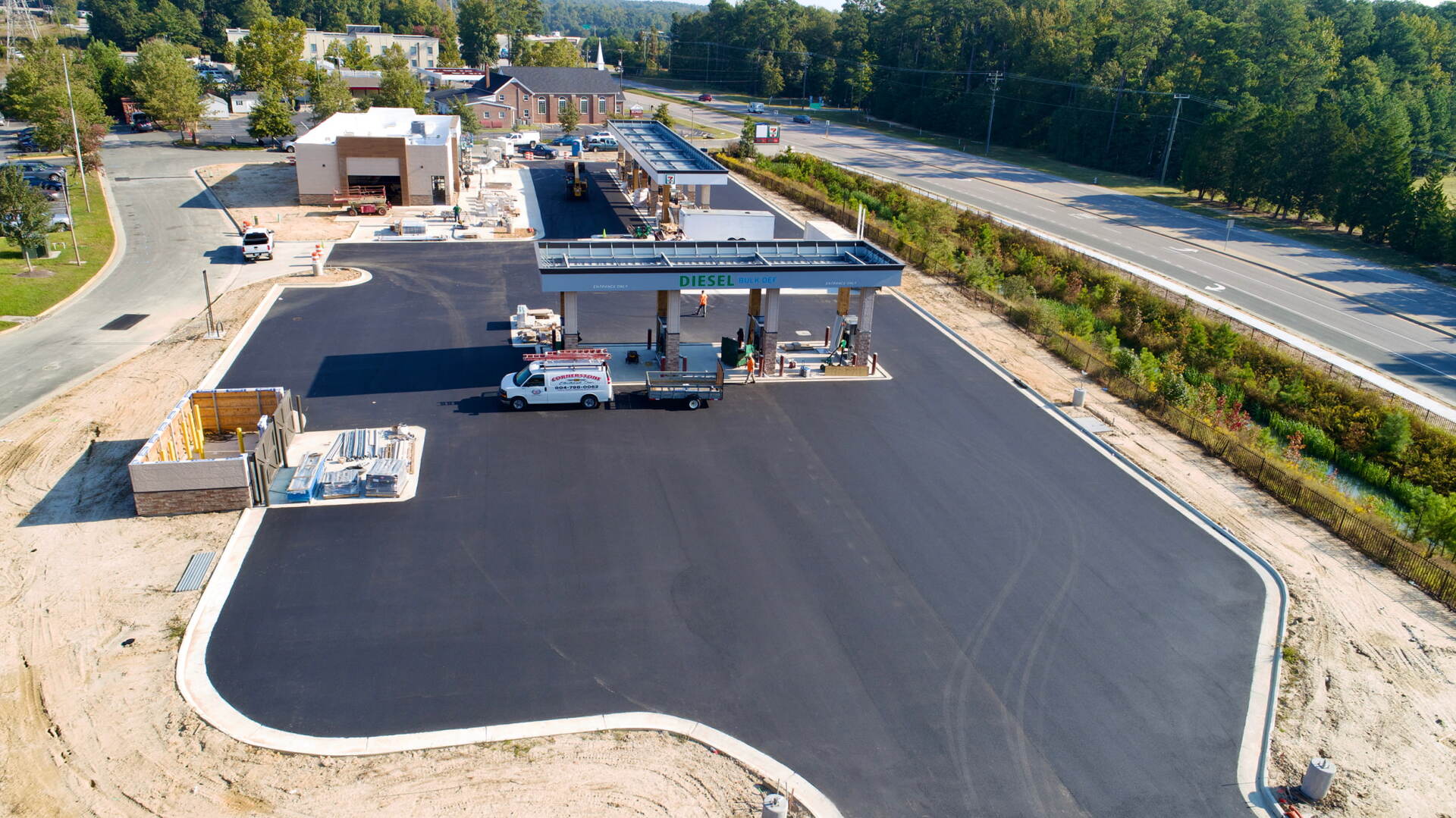 An aerial view of a newly paved gas station under construction, featuring a fuel canopy, parking lot, and nearby building.