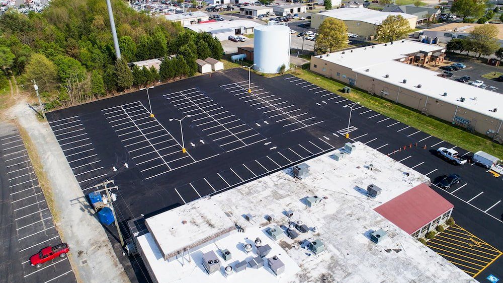 An aerial view of a freshly paved commercial parking lot with white striped spaces next to a large building and water tank.