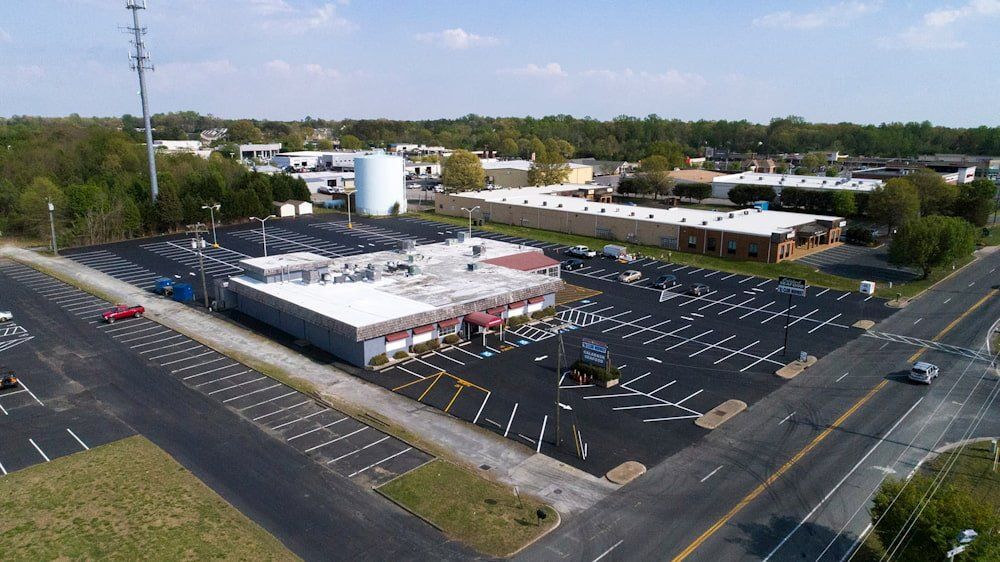 An aerial view of a building with a white roof and large parking lot situated next to a road and green space.
