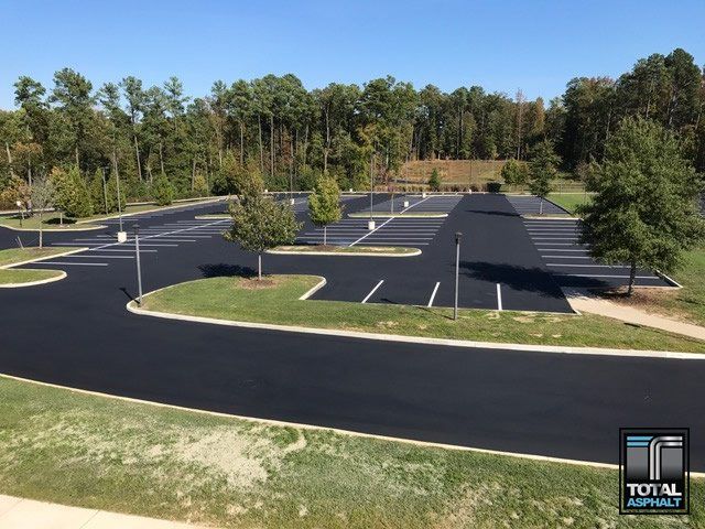 Newly paved dark asphalt parking lot with white-painted lines and surrounding grass, trees, and blue sky.