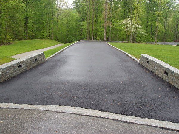 A new asphalt driveway entrance bordered by stone walls with integrated lighting, set against a green, wooded background.