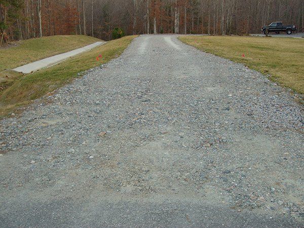 A gravel driveway leads from a paved road toward a house, flanked by grassy banks and a line of trees in the background.