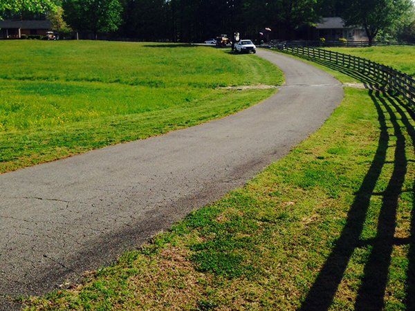 An asphalt driveway curves through a green, grassy field alongside a long wooden fence, leading toward a house in the back.