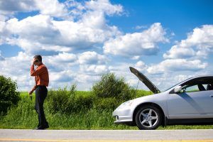 Man on phone next to a silver car with the hood up, roadside. Bright sunny day, blue sky, green field.