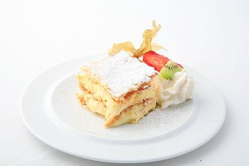 Fetta di torta a strati ricoperta di zucchero a velo, con fragole, kiwi e panna montata su un piatto bianco.