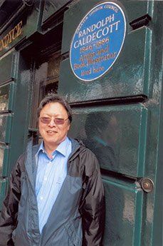 Man standing by a blue plaque honoring Randolph Caldecott. Dark green door, light blue shirt, and black jacket.