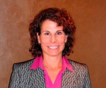 Dr. Elizabeth - Sacrey & Sacrey Dentistry in Geneva, IL Woman with curly brown hair smiling, wearing a pink shirt and patterned blazer, against a brown wall.