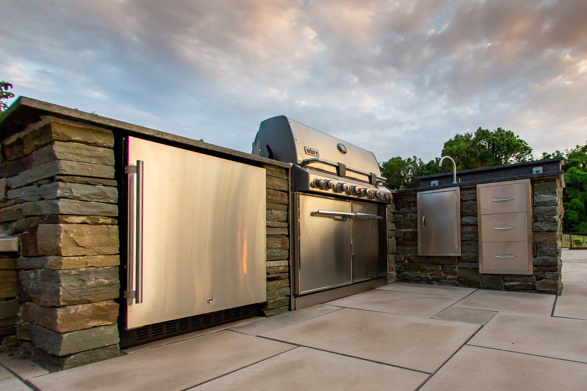 Outdoor kitchen with stainless steel appliances built into a stone structure on a stone patio.