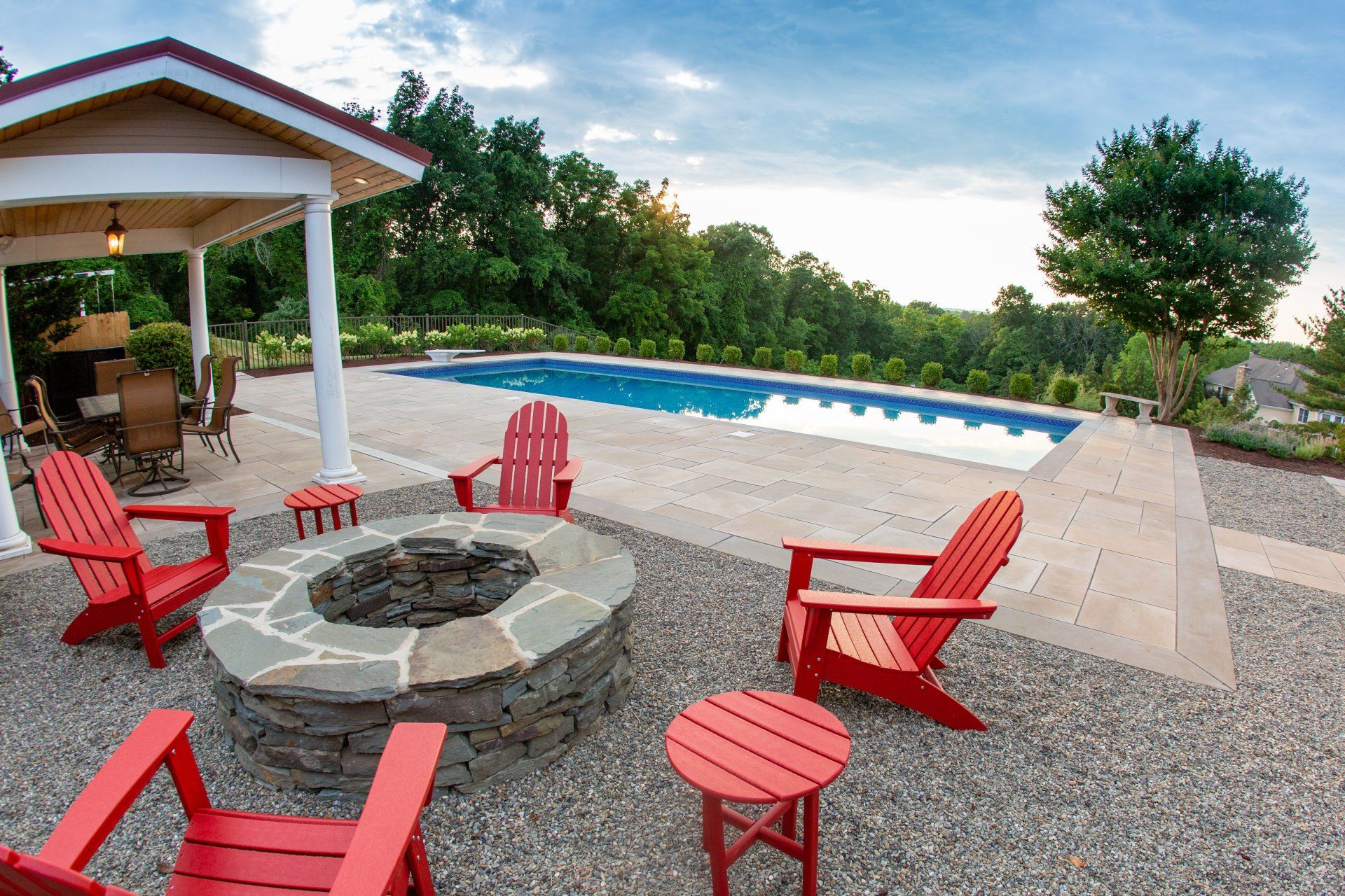 Red Adirondack chairs surround a stone fire pit near a pool and gazebo in an outdoor setting.