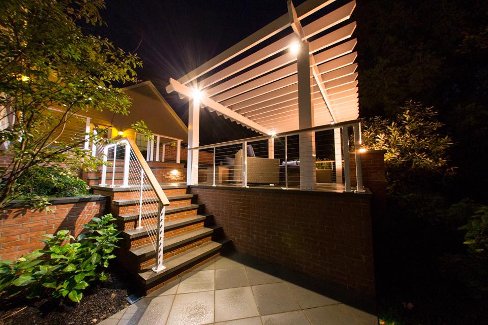 Night view of an outdoor patio with a pergola, brick walls, and steps, illuminated by lights.