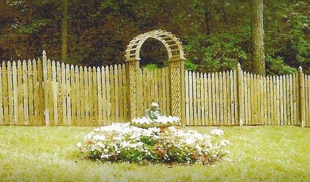 White picket fence with arched gate behind a flower bed in a sunlit garden clearing.
