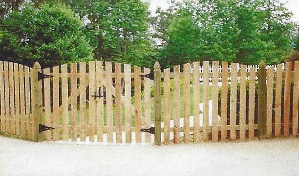 Wooden picket fence with curved gate in front of green trees and shrubbery