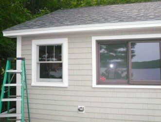 A side view of a house with light tan shingle siding, a dark roof, two windows with white trim, and a green step ladder.