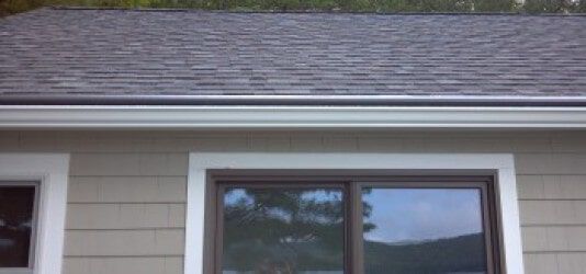 A tan house exterior with white window trim and a gutter installed below a shingled roof.