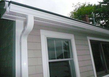 White gutter and downspout installed on the corner of a residential house with light-colored siding and window trim.