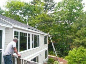 A person stands on a wooden deck looking at a house with light-colored siding and a ladder leaned against the roofline.