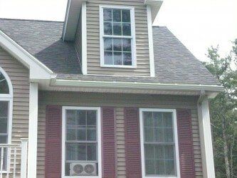 A tan-sided house with a dormer window, two ground-floor windows with red shutters, and a window air conditioning unit.