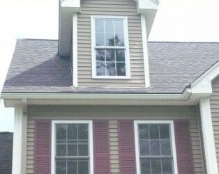 A tan-sided house exterior featuring a dormer window above two lower windows with red shutters under a grey shingled roof.