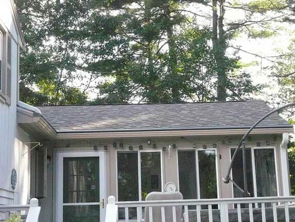 A light-colored house extension with a gray shingled roof, a glass door, and large windows, viewed from a wooden deck.