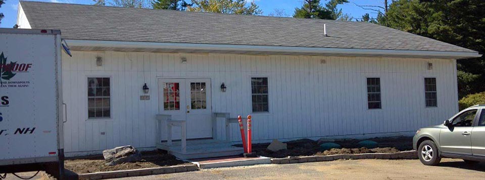 A single-story white building with a gray shingled roof, a central double door entrance, and a parking area in front.