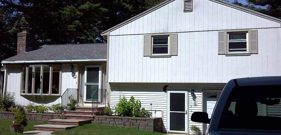 A white, two-story house with vertical siding, a bay window, and a stone retaining wall in the front yard.