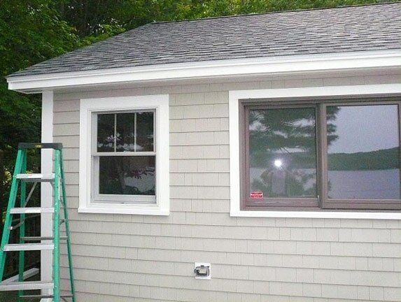 A green ladder stands against the side of a light gray shingled house with two windows and a roof overhang.