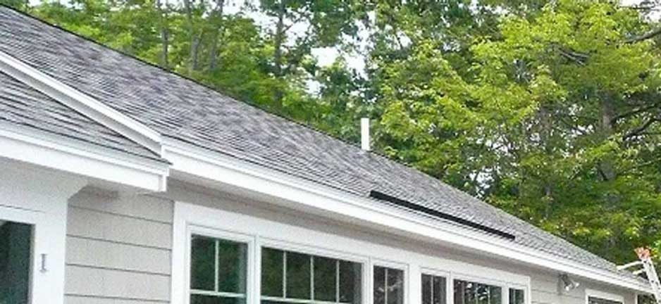 A gray-shingled house exterior with white trim, large windows, and a white vent pipe on a sloped roof near green trees.