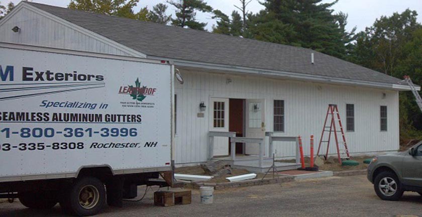 A white work truck parked outside a single-story white building, with a ladder leaning against the roof during construction.