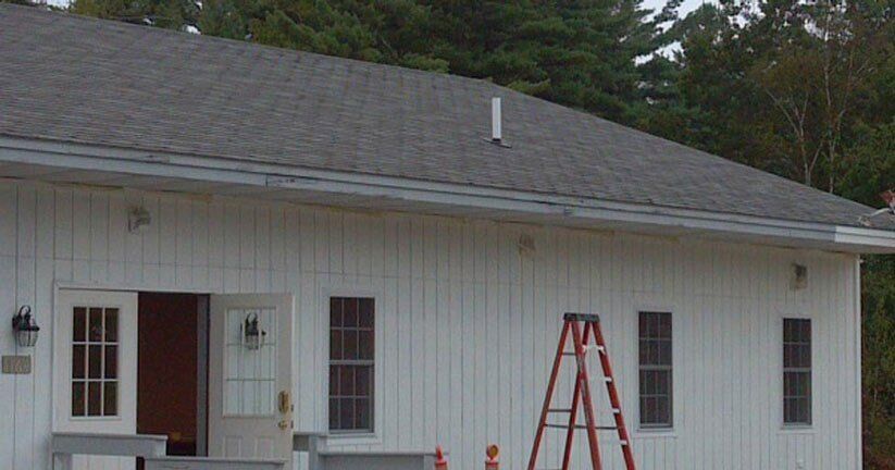 A white building with vertical siding, a grey shingled roof, a double door entrance, and a red ladder leaning against it.