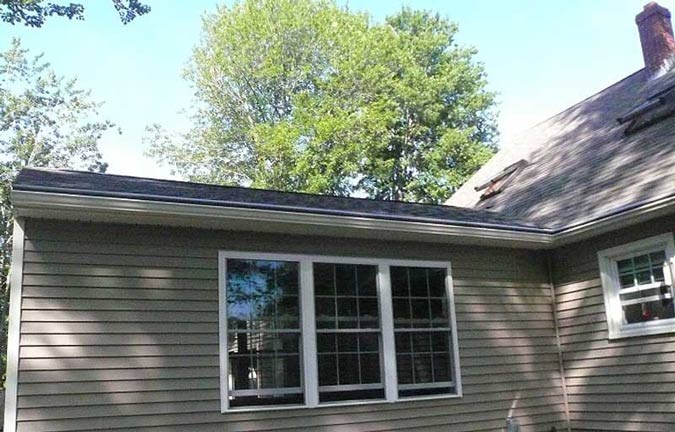 A side view of a house with light gray siding, a dark shingled roof, a three-panel window, and mature green trees above.