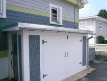 A shed or storage unit extension on the side of a house, featuring gray shingles, white doors, and a metal roof.