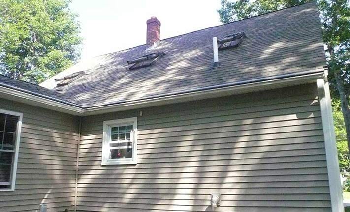 A tan-sided house exterior featuring an asphalt-shingle roof, a red brick chimney, two small roof vents, and a window.