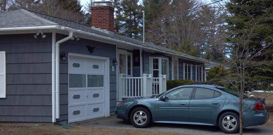 A grey, single-story house with a white garage door and a teal car parked in the driveway surrounded by trees.