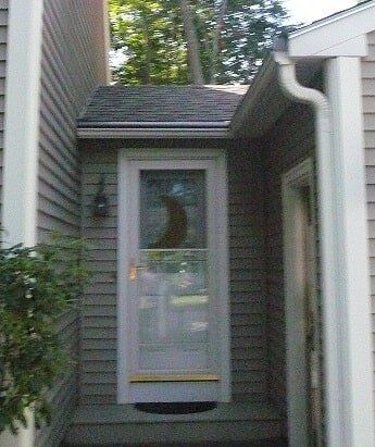 A white storm door with a crescent moon decorative insert, set in a gray siding entryway with a white downspout.