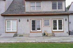 New red metal tile with white rain gutter - Seamless Gutter in Rochester, NH The rear exterior of a gray shingled house with three sets of glass doors, stone steps, and a large stone patio.