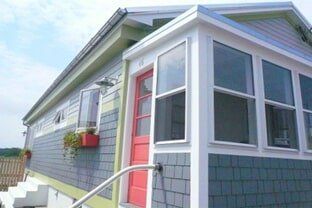 Rain gutter - Seamless Gutter in Rochester, NH Image A low-angle view of a cottage with light grey siding, a bright red door, and a white-framed glass sunroom addition.