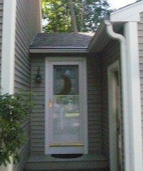 After Leaf Lock A white storm door with a crescent moon design, set in an entryway with grey siding and a small porch.