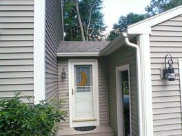 Before Leaf Lock A beige house entrance with a white storm door, a small roof overhang, two outdoor wall lamps, and surrounding greenery.