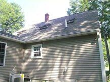 After Leaf Lock 2 A side view of a house exterior with light-colored vinyl siding, a roof with a brick chimney, two roof vents, and a window.