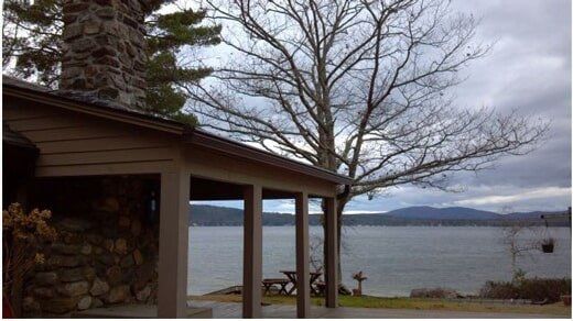 Without LeafLock — Seamless Gutters on a House in Portsmouth, NH A stone fireplace and wooden porch overlook a calm lake with mountains in the background under a cloudy sky.